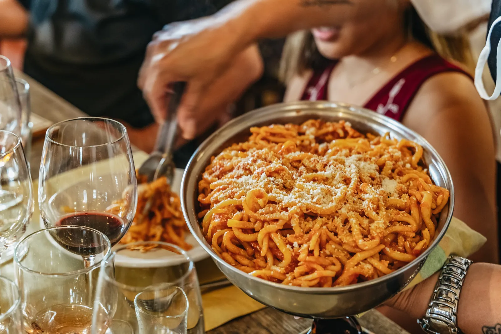 Steel dish of pici pasta with tomato sauce beside red and white wine glasses