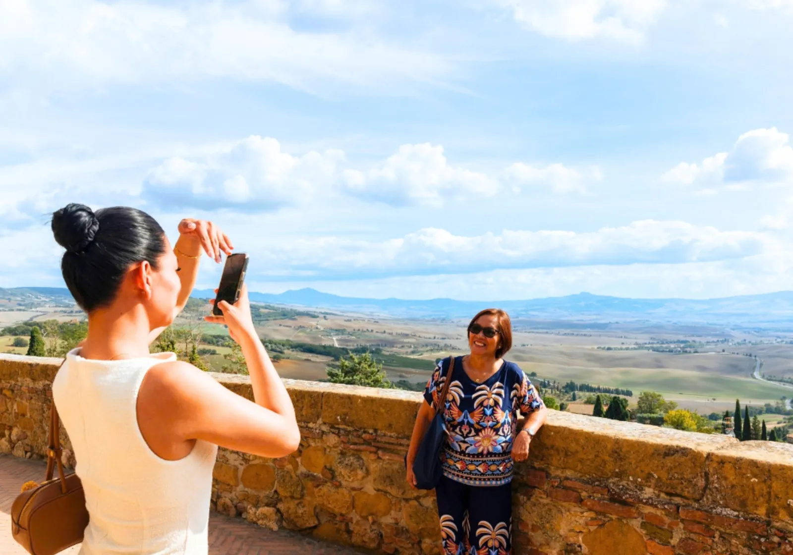 Visitor photographing a companion on a stone terrace wall above a wide Tuscan valley