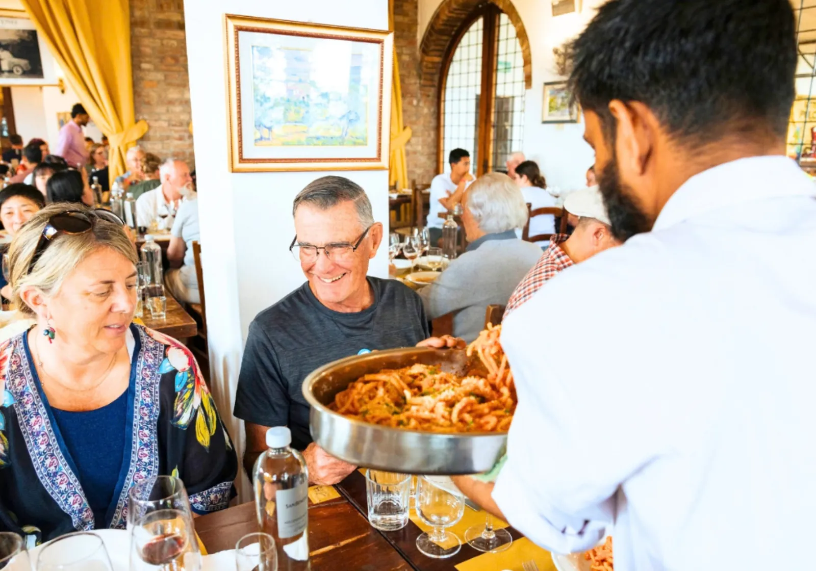 Waiter serving a steel dish of pasta to smiling diners in a busy restaurant