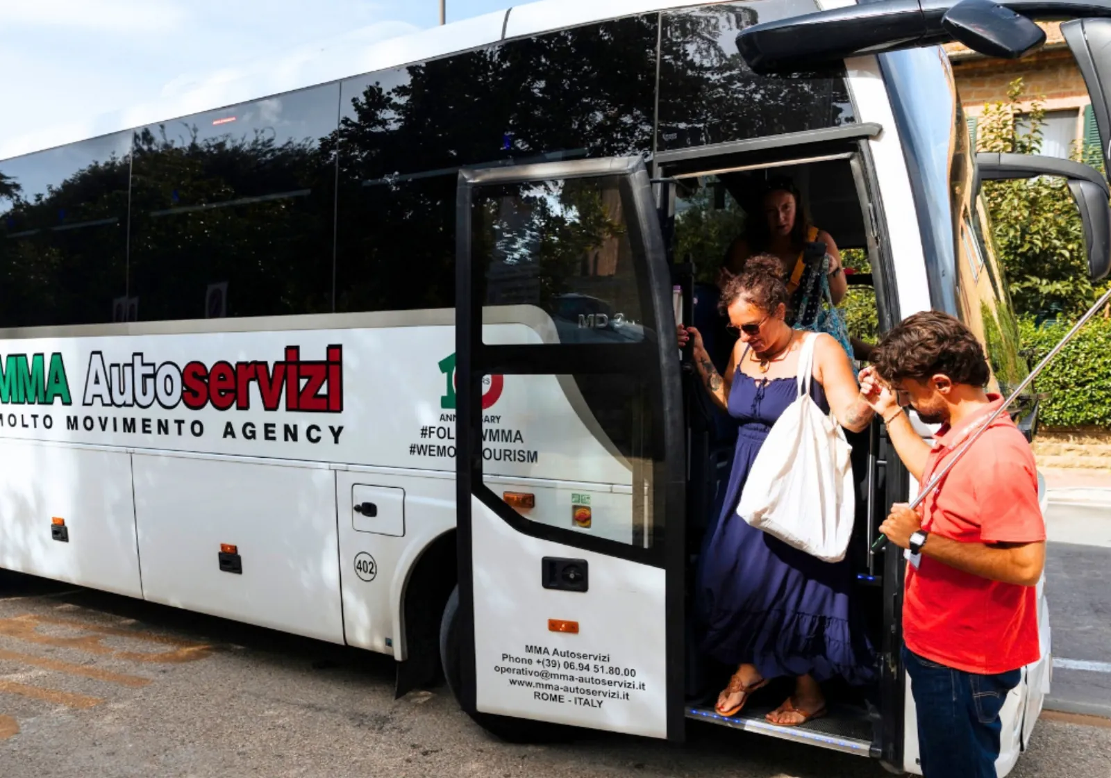 Passengers stepping off a white touring coach parked beside a stone wall