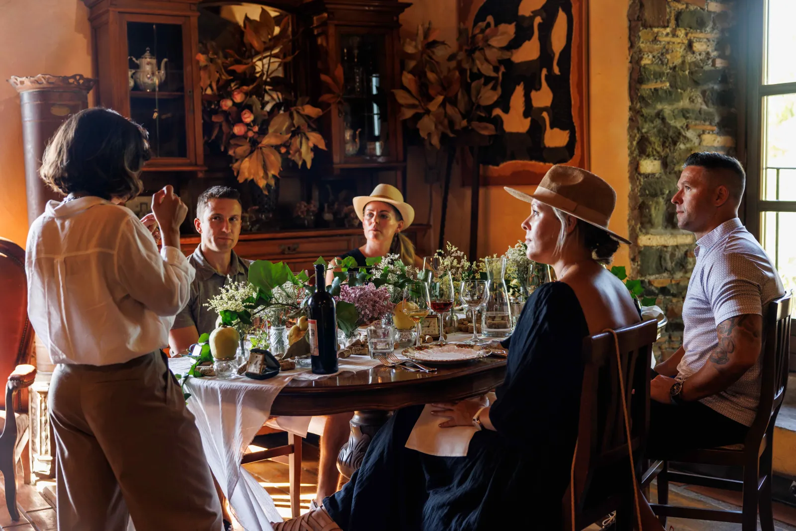 Guests seated around a flower-decorated table in a rustic dining room as a host speaks