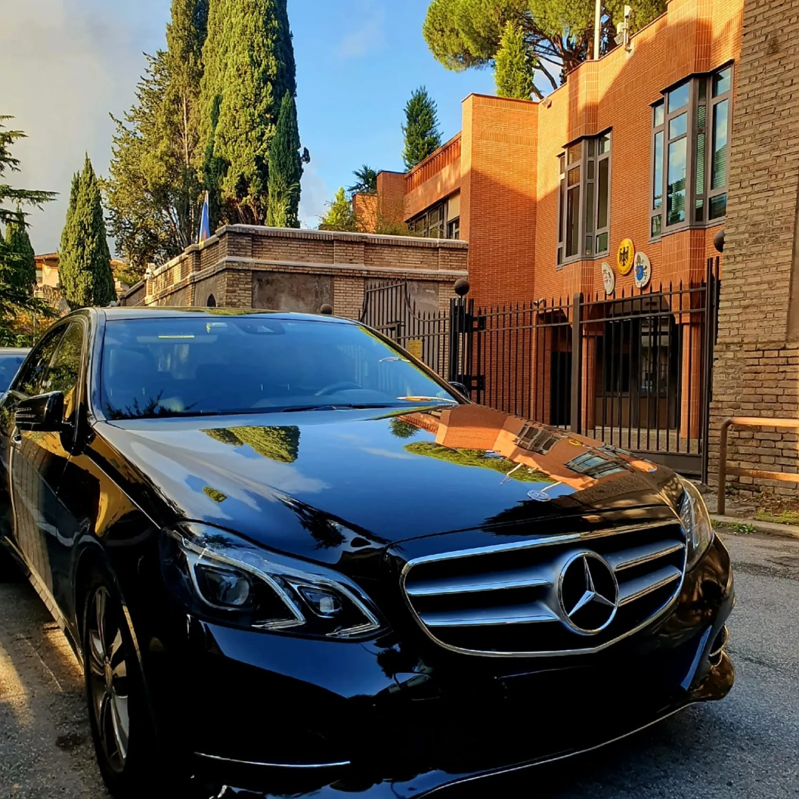 Black Mercedes sedan parked in front of a brick building and cypress trees