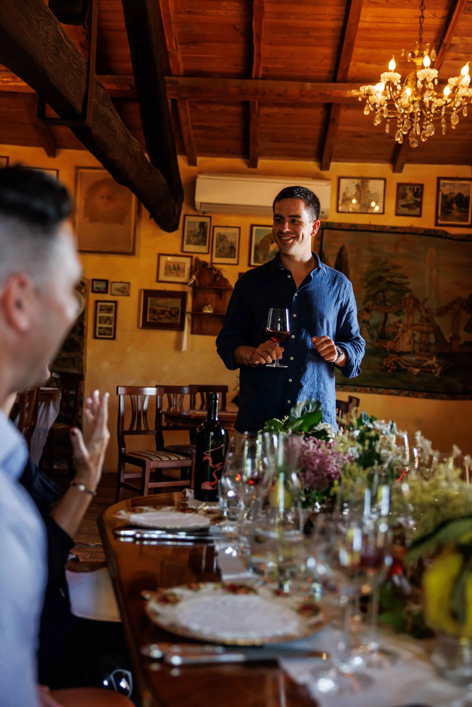Man holding a wine glass beside a long set table in a beam-ceilinged dining room