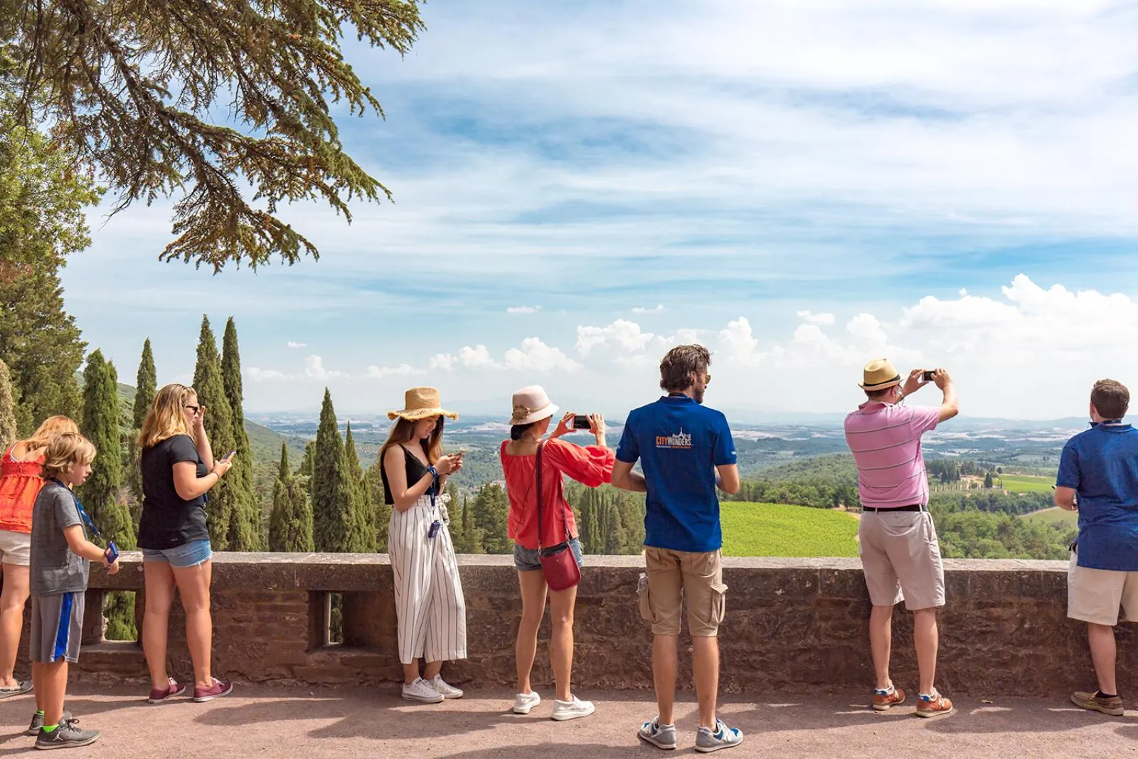 Visitors with phones along a stone terrace wall above a green valley with cypresses