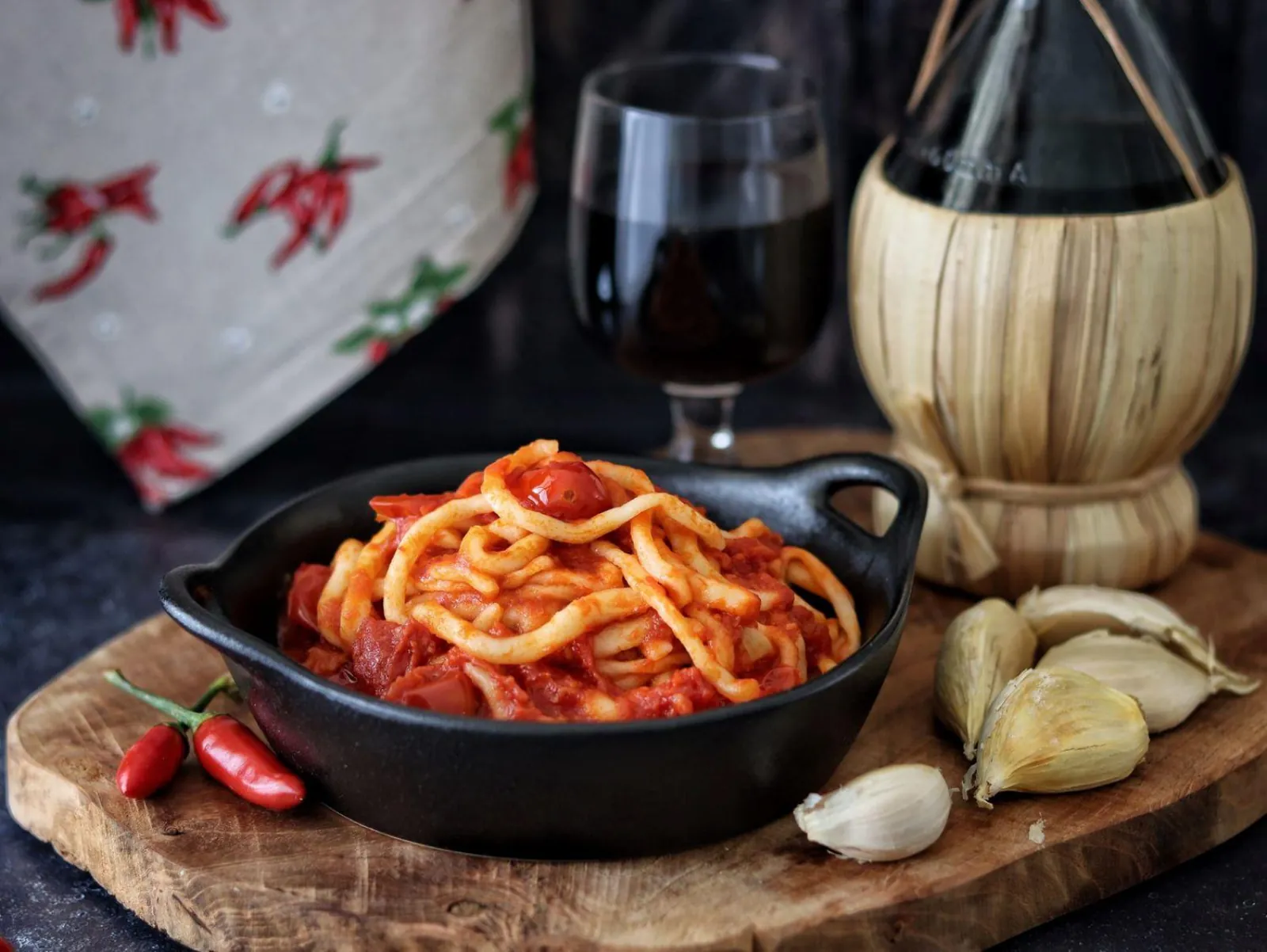 Dark skillet of spaghetti in tomato sauce with garlic, chilli peppers and a straw-covered wine flask