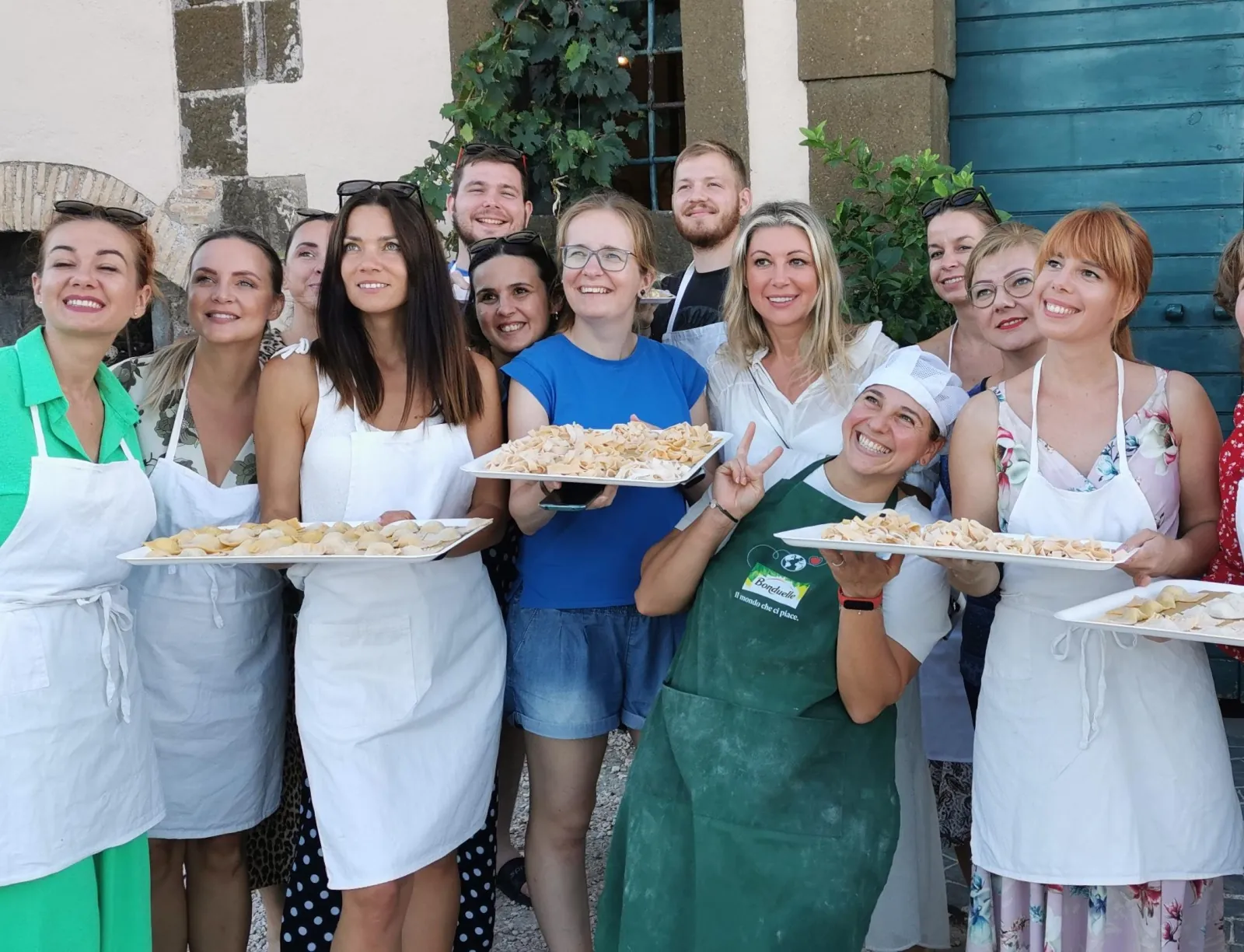 Group in aprons holding trays of fresh pasta in front of a farmhouse door