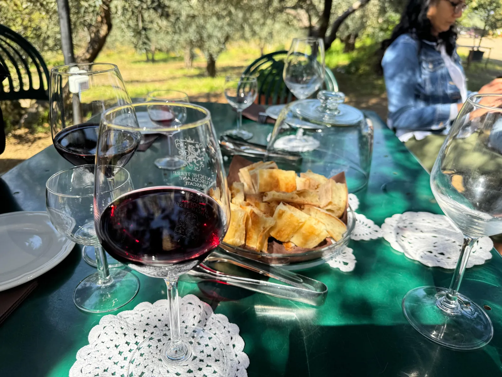 Red wine, flatbread crisps and wine glasses on a green table under olive trees