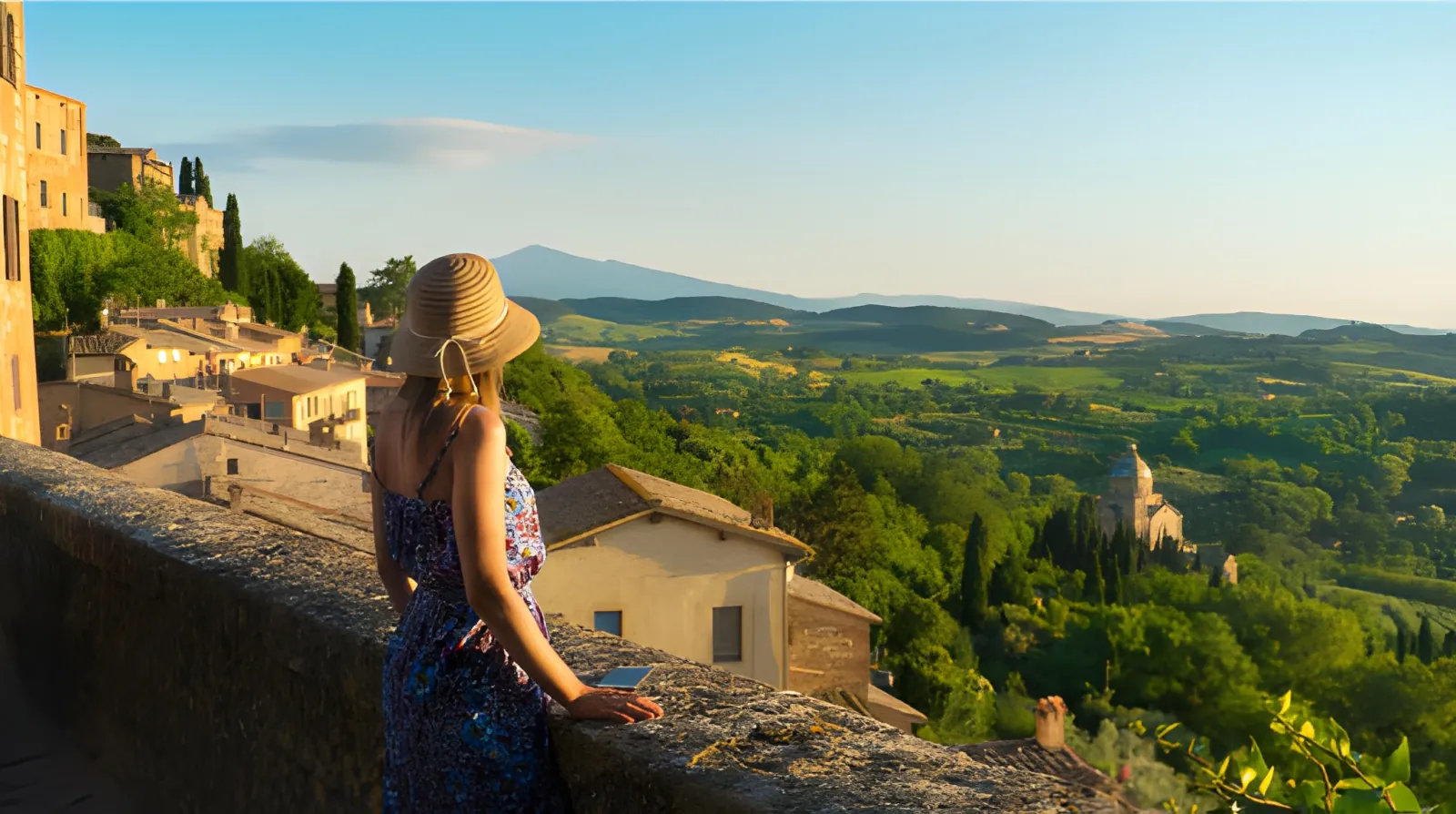 Woman in sun hat at a stone wall overlooking green Tuscan countryside and a church