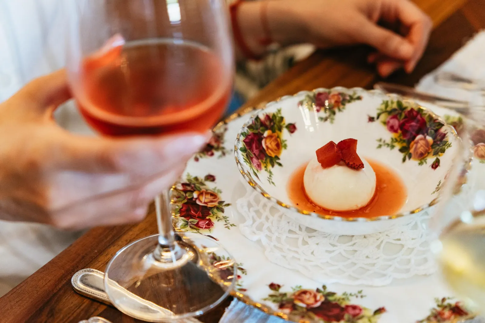 Hand holding a glass of rosé beside a floral bowl with a white dessert