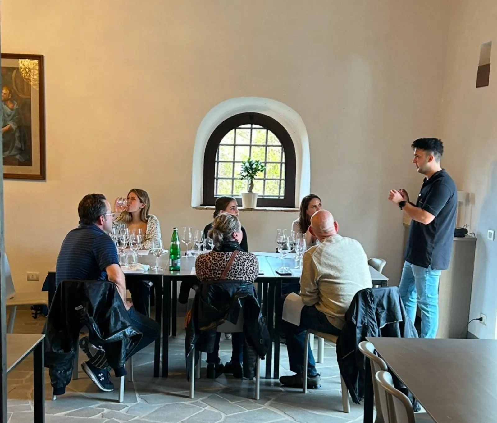 Small group around a table with wine glasses as a host speaks in a whitewashed room